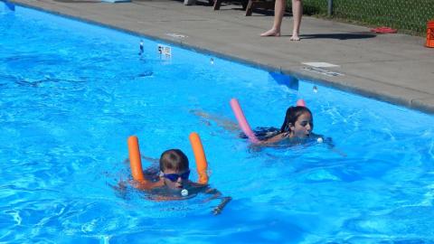 Campers swimming with pool noodles blowing ping pong balls across the pool during a race.