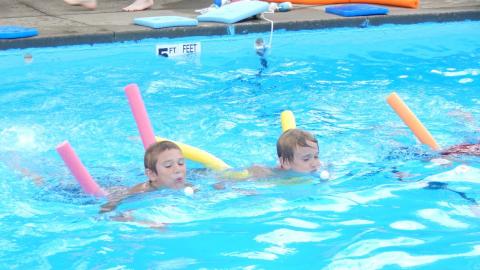 Campers swimming with pool noodles blowing ping pong balls across the pool during a race.