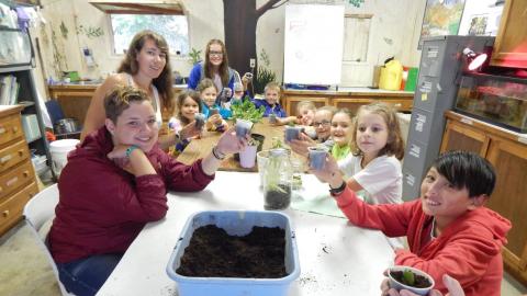 Campers seated at a table holding up plants they have potted to the camera and smiling.