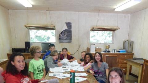 Kids seated around a table looking through old camp newspapers and smiling at the camera.