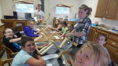 Youth sitting around a table string beads onto wooden looms looking at camera and smiling.