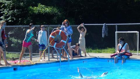 Youth throwing a rescue ring to pretend victim flailing in the pool as other campers watch from pool deck.
