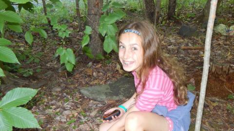 Camper in wooded are bending down to collect a geocache with GPS in their hand smiling at camera.