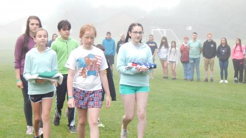 Colorguard walks toward the flagpole holding American and 4-H flags through circle of other campers.