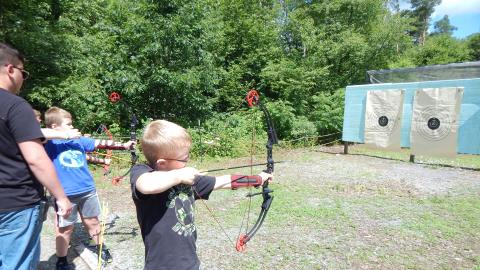 Youth standing on line with bows drawn and ready to fire as a counselor looks on.