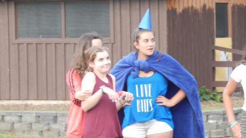 Three youth in a skit. One wears a blanket as a cape and cone on their head.