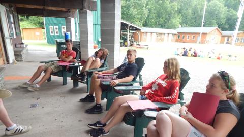 Teens sit in Adirondack chairs on the dining hall porch with folders listening to CIT Director.