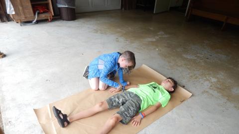 Camper lays on long roll of brown paper while other camper traces their outline.