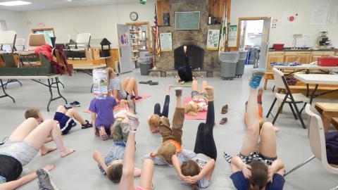 Campers lay on dining hall floor with hands behind head a feet raised up in the air following the lead of a counselor.