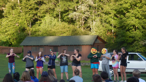 Cabin group stands together in front of camp audience making hearts with their hands. Two youth have emoji pillows in their sweatshirts as if they were their faces. 