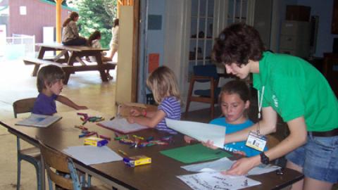 Day Camp Director works with three youth coloring at a table in the Day Camp room.