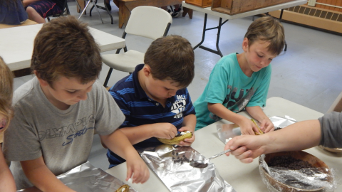 youth putting marshmallows and chocolate chips into a sliced banana over tin foil while seated around a table.