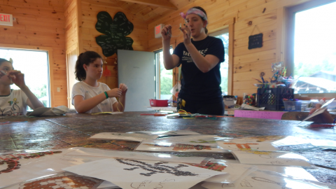 Craft director demonstrates beading technique to a group of campers seated around the table. Patterns they can choose to make are scattered across the table including a skunk, football, and many more.