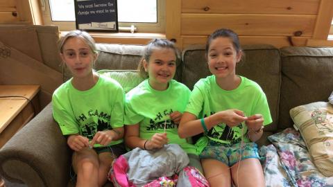 Three youth in matching camp t-shirts sit on the craft hall couch making boondoggles.
