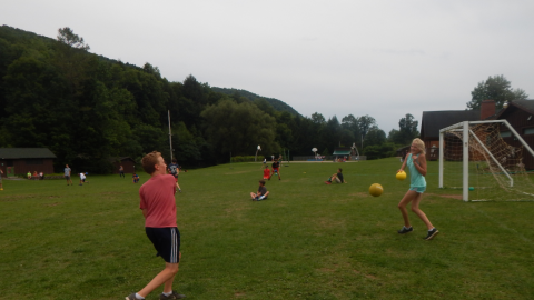Two youth run opposite each other holding foam balls while other campers are scattered in the background.