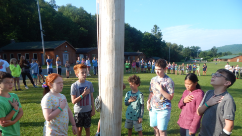 Group of youth saying the pledge of allegiance at the flagpole while the rest of camp follows along from a larger circle.