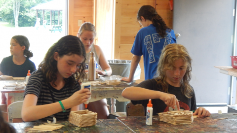 Campers seated at table in craft hall constructing boxes with glue and Popsicle sticks.