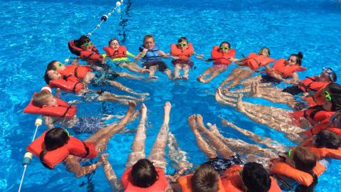 Group of youth in the pool wearing life jackets and linking arms to form an orange rescue circle.