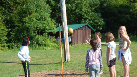 Five youth around a tether ball poll. Three watch while two are playing.