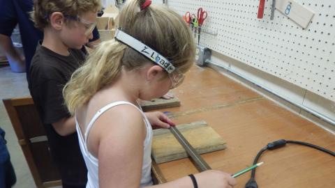 Youth wearing safety goggles measures a wooden board while holding a pencil.