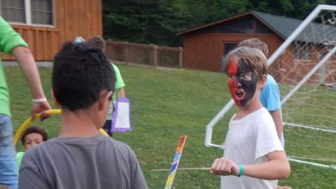 Youth engaged in wooden sword fight. Youth facing the camera has face fully painted with mouth open.