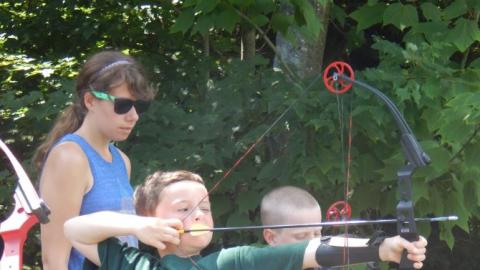 Youth standing on line with bows drawn ready to fire as counselor looks over their shoulders.