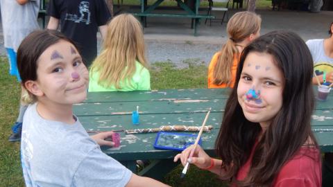 Two youth looking into camera with painted faces. One holds out their arm to the other who is holding a paintbrush.