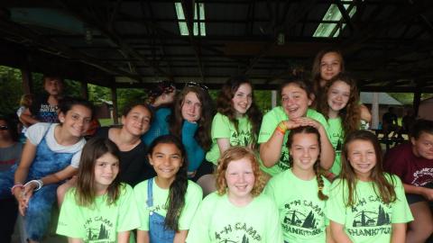 Medium group of youth wearing wearing camp t-shirts seated in the rec hall pavilion and smiling together at the camera.