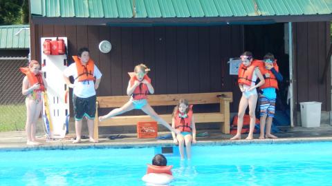 Camper jumping into pool toward counselor while wearing lifejacket with legs spread as other campers watch from the pool deck.
