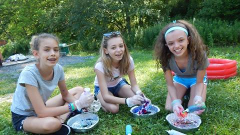 Three youth seated on grass wearing gloves and pouring die into tin pans smiling at camera.