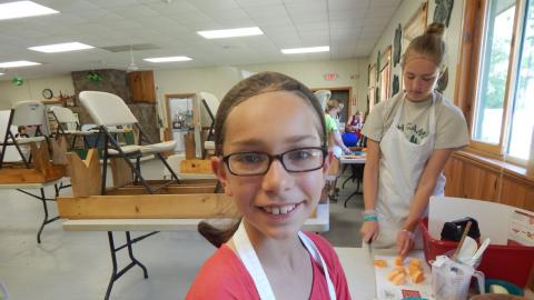 Youth smiling into camera wearing apron with measuring tools on counter and other youth chopping veggies in the background.