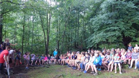 campers seated on benches in a wooded area watching a performance by a platoon group in front.