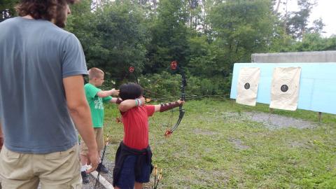 Youth standing on line with bows drawn aiming at paper targets as counselor looks on.
