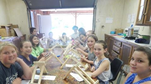 Youth seated around a table stringing beads onto wooden looms and all looking into the camera smiling.