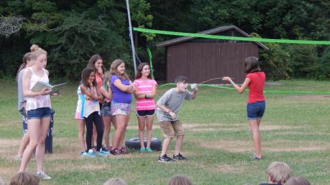 Cabin group standing together behind one camper who speaks into a cup on the end of a stick held by another camper as counselor to the side reads a story.