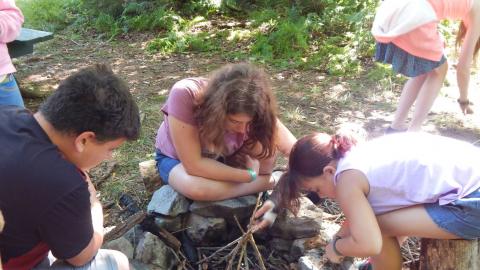 Youth lean over a fire pit to construct a teepee.
