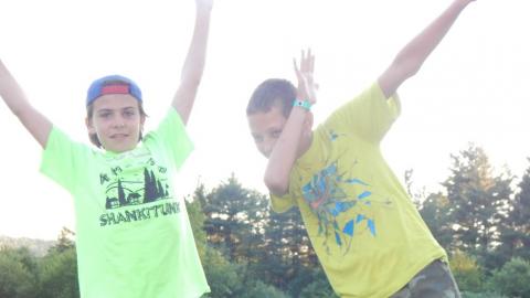 Two youth in foreground looking at camera with arms raised overhead to the side.Rest of camp in background seated on lawn.