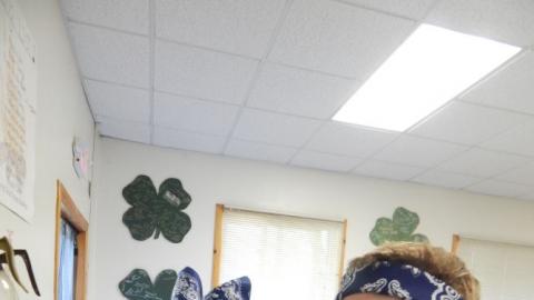 Five youth lined up at a counter wearing bandannas and smiling at the camera as they work with food processors. The youth closest to the camera points both hands at the camera.