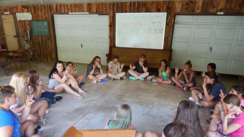 Youth in large circle on the floor doing sign language led by a counselor.