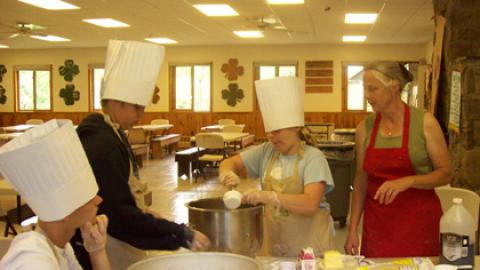 three youth in paper chef hats work under the supervision of CCE Educator in chefs club to add ingredients to a large metal pot.