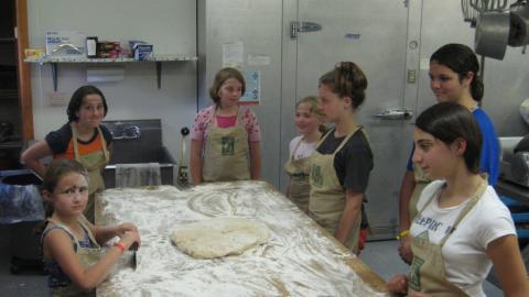 youth in Chef's Club stand around a kitchen counter covered in flour and dough with half a tray of shaped garlic knots.