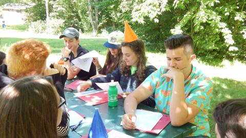 Youth seated at picnic table draw on paper shield. One youth is wearing cone hat.
