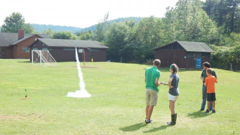 Two campers stand with counselors as their model rockets launch into the air.