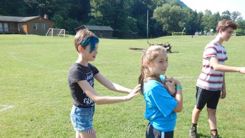 One camper looks toward the camera with rms crossed in front of them as their partner braces on dominate leg with arms outstretched to catch them in a trust fall.