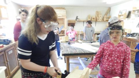 Youth using a jig while they saw into a board with a handsaw while another camper looks on. A counselor in the background looks around the room at other youth working.
