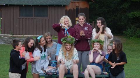 Cabin group on and around a picnic table smiling at counselors who are wearing crowns of vines.