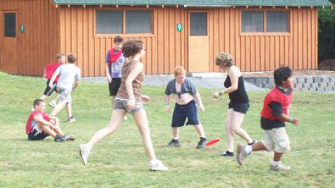 eight youth engaged in a game. Half the youth are wearing red pinnies. Youth are running across a line of frisbees to mark center field.