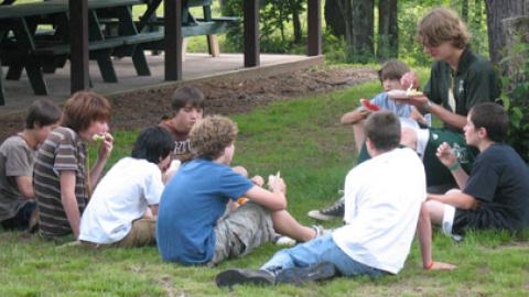 Group of campers sit together with a counselor eating dinner near a tree.