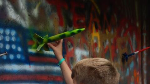Youth reaches for a rocket painted green with dark spots from a wall of rockets out behind the science center.