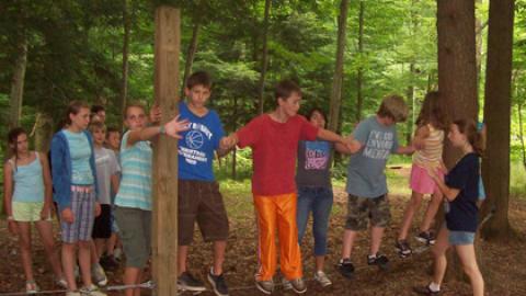 group of campers holding hands suspended on a cable a few inches from the ground bolted into neighboring trees and posts.